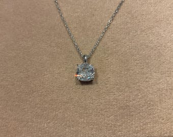 May include: A silver necklace with a square, cushion-cut diamond pendant. The diamond sparkles, reflecting light. The chain is delicate and hangs on a neutral-colored surface. A piece of jewelry.