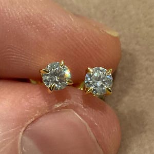 May include: A pair of gold stud earrings, each featuring a round, clear gemstone. The gemstones are held in place by four gold prongs. The earrings are shown against a neutral background.