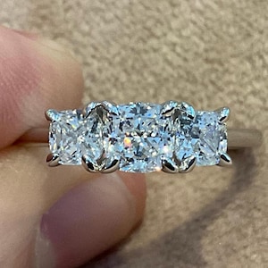 May include: A silver ring featuring three square-cut, clear gemstones. The ring is held up, displaying the sparkling stones. The ring is a symbol of commitment and love. The gemstones are set in a silver band.