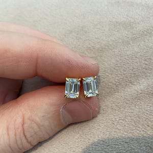 May include: A pair of gold stud earrings, each featuring a rectangular, clear gemstone. The earrings are held between fingers against a neutral background. The gemstones have a faceted cut, reflecting light.