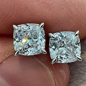Puede incluir: Un par de pendientes de diamantes de talla cuadrada. Cada pendiente presenta un diamante grande, claro y brillante engastado en una montura de garras de plata u oro blanco. Los diamantes tienen un corte cojín y reflejan la luz, creando una exhibición brillante.