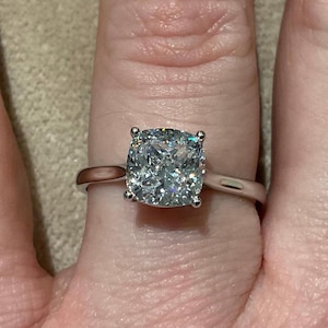 May include: A close-up of a silver ring with a large, square-cut, clear gemstone. The gemstone is held in place by four prongs. The ring is on a finger.