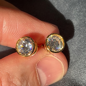 Könnte beinhalten: Ein Paar goldfarbene Ohrstecker, jeweils mit einem großen, runden, klaren Edelstein. Die Edelsteine sind in einer Lünettenfassung gefasst. Die Ohrringe werden zwischen den Fingern vor einem dunklen Hintergrund gehalten.
