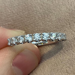 May include: A silver ring with a continuous row of round, clear gemstones. The ring is held by a person's fingers against a neutral background. The gemstones are evenly spaced and appear to be set in a channel or shared-prong setting.