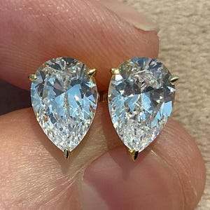 May include: A pair of pear-shaped diamond stud earrings. The diamonds are clear and faceted, set in gold prongs. The earrings are displayed against a neutral background.