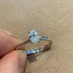 Op de afbeelding: Een zilveren ring met een grote, ovaal geslepen diamant. De diamant is het middelpunt, gezet op een eenvoudige band. De ring wordt door een hand vastgehouden, wat het ontwerp en de schittering benadrukt. De ring is een sieraad.