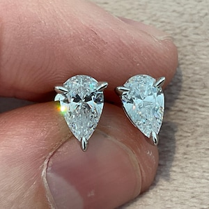 Puede incluir: Un par de pendientes de diamantes con forma de pera. Los diamantes claros y brillantes están engastados en metal plateado. Los pendientes se sostienen entre los dedos sobre un fondo neutro.