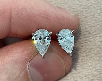 May include: A pair of pear-shaped diamond stud earrings. The diamonds are clear and sparkling, set in a silver metal. The earrings are held between fingers against a neutral background.