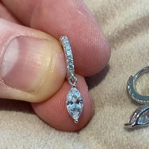 Puede incluir: Un pendiente de plata con un colgante de piedra preciosa con corte marquesa. El pendiente presenta un pequeño aro con piedras claras incrustadas. Un segundo pendiente es visible en el fondo.