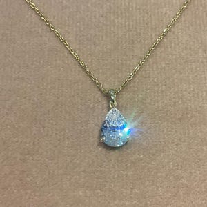 Peut inclure: Un collier doré avec un pendentif en forme de poire. Le pendentif est une pierre transparente et étincelante avec un effet irisé bleu et vert. Le collier est présenté sur un fond neutre.