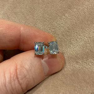 May include: A pair of square-cut, clear gemstone stud earrings. The earrings are held between fingers against a neutral background. The gemstones reflect light, creating a sparkling effect. The earrings are set in a silver or white metal.