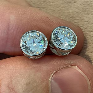 May include: Two round stud earrings with a bezel setting. Each earring has a large, clear, faceted gemstone set in a silver-coloured metal. The gemstones sparkle with internal reflections, creating a brilliant display.