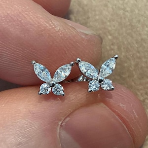 May include: A pair of silver butterfly stud earrings. Each earring features two marquise-cut and two round-cut clear gemstones, arranged to resemble a butterfly. The earrings are displayed against a blurred background.