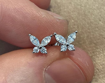May include: A pair of silver butterfly stud earrings. Each earring features two marquise-cut and two round-cut clear gemstones, arranged to resemble a butterfly. The earrings are displayed against a blurred background.