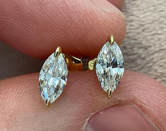 May include: A pair of marquise-cut diamond stud earrings. The diamonds are set in a gold bezel setting. The earrings are held between fingers. The diamonds are clear and sparkling, reflecting light.