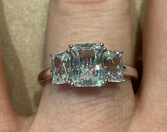 May include: A silver ring with three large, clear, rectangular-cut stones. The center stone is slightly larger than the two side stones. The ring is set on a finger, showcasing the sparkling facets of the gems.