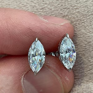 May include: A pair of marquise-cut stud earrings. Each earring features a clear, faceted gemstone set in a silver-colored metal. The gemstones have a brilliant sparkle, reflecting light. The earrings are displayed against a neutral background.