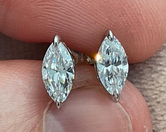May include: A pair of marquise-cut diamond stud earrings. The diamonds are set in a silver metal, and the earrings are held between fingers. The diamonds are clear and reflect light.