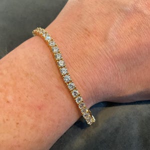 Puede incluir: Un brazalete dorado con una línea de piedras preciosas redondas y transparentes. El brazalete se lleva en la muñeca. Las piedras preciosas están espaciadas uniformemente y reflejan la luz. El engaste dorado es visible.