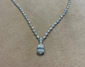 May include: Elegant necklace featuring a delicate chain adorned with numerous small, sparkling stones. The focal point is a pear-shaped, brilliant-cut gemstone suspended from a cluster of smaller stones. The necklace is displayed on a neutral, textured surface.