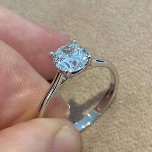 Puede incluir: Primer plano de un anillo de plata con una gran gema redonda y transparente. El anillo tiene un diseño simple y elegante con una banda delgada y cuatro garras que sujetan la gema. El anillo está sostenido por dedos.