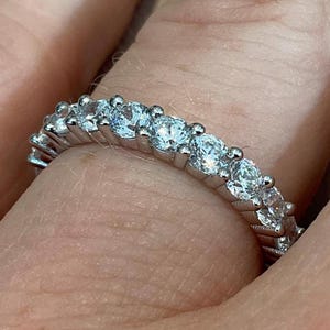 May include: A silver ring featuring a continuous row of clear, round gemstones. The ring is set with multiple small, sparkling stones, creating a band of light. The ring is shown on a finger.