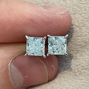 May include: A pair of square-cut diamond stud earrings with four-prong settings. The diamonds are clear and sparkling, set in a silver-coloured metal. The earrings are shown against a neutral background.