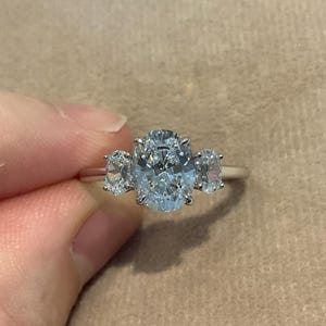 Könnte beinhalten: Ein silberner Ring mit einem großen, oval geschliffenen Mittelstein, flankiert von zwei kleineren, oval geschliffenen Steinen. Der Ring ist auf einem silbernen Band gefasst, und die Edelsteine funkeln. Der Ring wird vor einem neutralen Hintergrund gehalten.