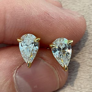 Op de afbeelding: Een paar peervormige diamantstud-oorbellen. De heldere, facetgeslepen diamanten zijn gezet in gouden prongen. De oorbellen worden tussen de vingers gehouden, wat hun glans en ontwerp benadrukt. Een klassiek sieraad.