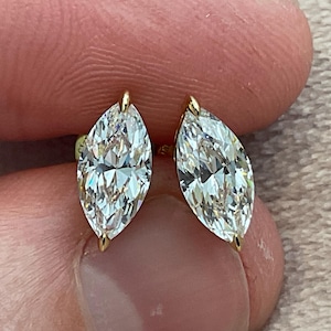 May include: A pair of marquise-cut diamond stud earrings. The diamonds are set in gold prongs, and the earrings are held between fingers. The diamonds have a brilliant cut, reflecting light. The earrings are a classic jewelry item.
