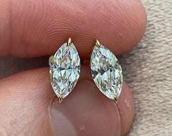 May include: A pair of marquise-cut diamond stud earrings. The diamonds are set in gold prongs, and the earrings are held between fingers. The diamonds have a brilliant cut, reflecting light. The earrings are a classic jewelry item.