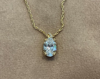 May include: A gold-colored necklace with a large, oval-shaped, clear gemstone pendant. The pendant is set in a gold-colored metal and hangs from a delicate gold chain. The gemstone reflects light, creating a sparkling effect.
