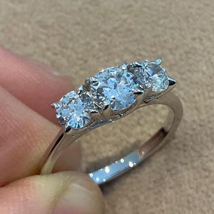 Könnte beinhalten: Ein silberner Ring mit drei runden, klaren Edelsteinen. Die Edelsteine sind in einer Reihe auf einem polierten Silberband angeordnet. Der Ring wird vor einem neutralen Hintergrund gehalten, um das Funkeln der Steine hervorzuheben.