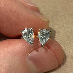 May include: A pair of diamond stud earrings. Each earring has a pear-shaped, clear diamond set in a silver or white gold prong setting. The earrings are held between fingers against a neutral background.