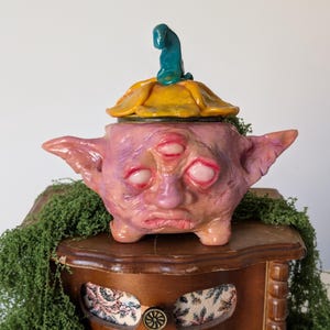 以下が含まれることがあります： 手作りのセラミック容器と蓋。容器は薄紫色で、3つの目、小さな耳、下向きの口があります。蓋は黄色で、上にターコイズの彫刻が施されています。容器は木製の表面に置かれています。