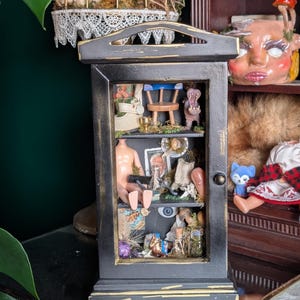 May include: A black wooden cabinet with distressed gold edges featuring two shelves filled with miniature objects including a doll torso, a small blue chair, a hippopotamus figurine, glass bottles, moss, and various small trinkets arranged inside.