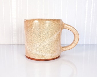 Op de afbeelding: Een beige keramische mok met een gespikkeld glazuur en een afgeronde handgreep. De mok heeft een licht getextureerd oppervlak en een lichtbruine rand.