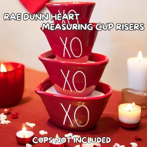 May include: Three red ceramic measuring cup risers in the shape of hearts with the word "XO" on each. The risers are stacked on top of each other. The text "RAE DUNN HEART MEASURING CUP RISERS" is at the top of the image. The text "CUPS NOT INCLUDED" is at the bottom of the image.