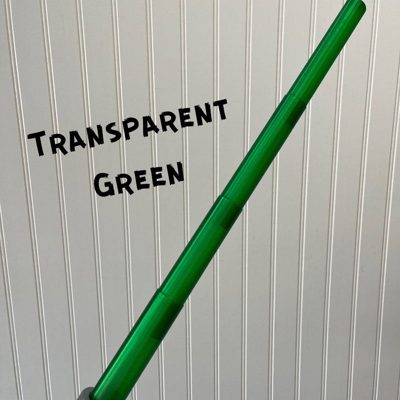 Puede incluir: Un accesorio de sable de luz verde transparente. El sable de luz es de pl&aacute;stico y extensible. El texto "TRANSPARENT GREEN" est&aacute; escrito sobre un fondo blanco detr&aacute;s del sable de luz.
