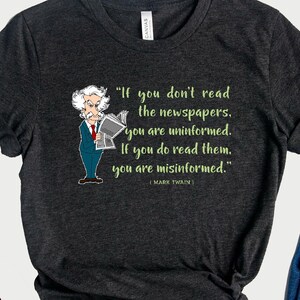 May include: A dark gray t-shirt with a quote by Mark Twain that reads 'If you don't read the newspapers, you are uninformed. If you do read them, you are misinformed.' The quote is printed in green text and is accompanied by a cartoon illustration of Mark Twain reading a newspaper. A black and yellow logo with the words 'Crazy Cats Merch' is in the bottom left corner of the image.
