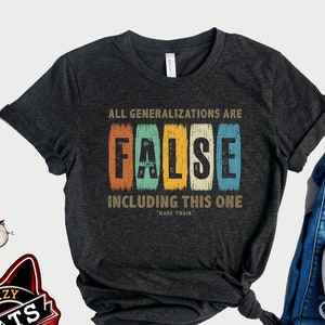 May include: A dark gray t-shirt with a vintage-style graphic that reads "All generalizations are false, including this one." The graphic is in a rainbow color scheme and includes a quote from Mark Twain. The t-shirt is laid out on a white surface with a pair of jeans and a camera.