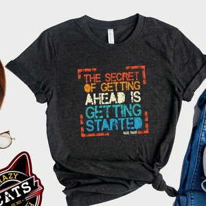 May include: A dark gray t-shirt with a vintage-style graphic that reads "The Secret of Getting Ahead is Getting Started - Mark Twain".