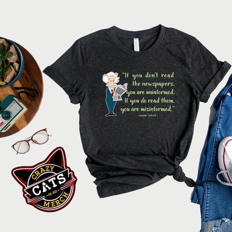 May include: A dark gray t-shirt with a quote by Mark Twain that reads 'If you don't read the newspapers, you are uninformed. If you do read them, you are misinformed.' The quote is printed in green text and is accompanied by a cartoon illustration of Mark Twain reading a newspaper. A black and yellow logo with the words 'Crazy Cats Merch' is in the bottom left corner of the image.