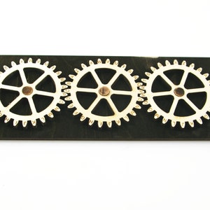 Può includere: Tre ingranaggi in legno con l'alfabeto stampato su di essi, montati su una tavola di legno marrone scuro. Gli ingranaggi sono bianchi e hanno un centro color rame.