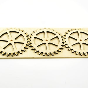Può includere: Tre ingranaggi in legno con l'alfabeto stampato sul bordo esterno. Gli ingranaggi sono montati su una base in legno.