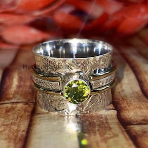 May include: A silver-colored spinner ring with a central, round, light green gemstone. The ring has a textured, floral design and gold-colored bands. The background is a wooden surface with red and orange floral accents.