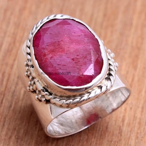 May include: A silver ring with a large, oval-cut ruby gemstone. The ruby is a deep red colour. The ring has a textured, rope-like border around the gemstone and a wide band. The text "Muskan Handcraft" is visible.