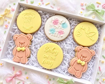 May include: A white gift box containing five decorated cookies: two light pink teddy bears with yellow bow ties, two yellow circular cookies featuring a baby onesie and a stork, and one white circular cookie with the text He or She can't wait to SEE! and Baby ON THE way.