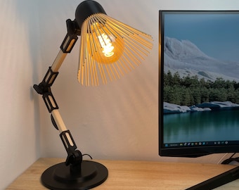 May include: A black desk lamp with a wooden shade and a light bulb turned on. The lamp is on a wooden desk with a notebook, pen, computer monitor, and keyboard.
