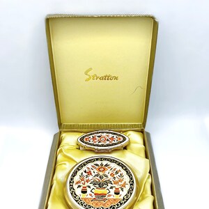 Peut inclure: Un miroir compact doré et un petit compact assorti dans une boîte doublée de satin jaune. Les deux compacts présentent un motif floral avec des accents orange, rouge et noir. La boîte est étiquetée "Stratton".